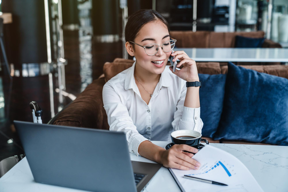 Asian businesswoman talking on the phone while sitting at a table, discussing website cost options for 2024.