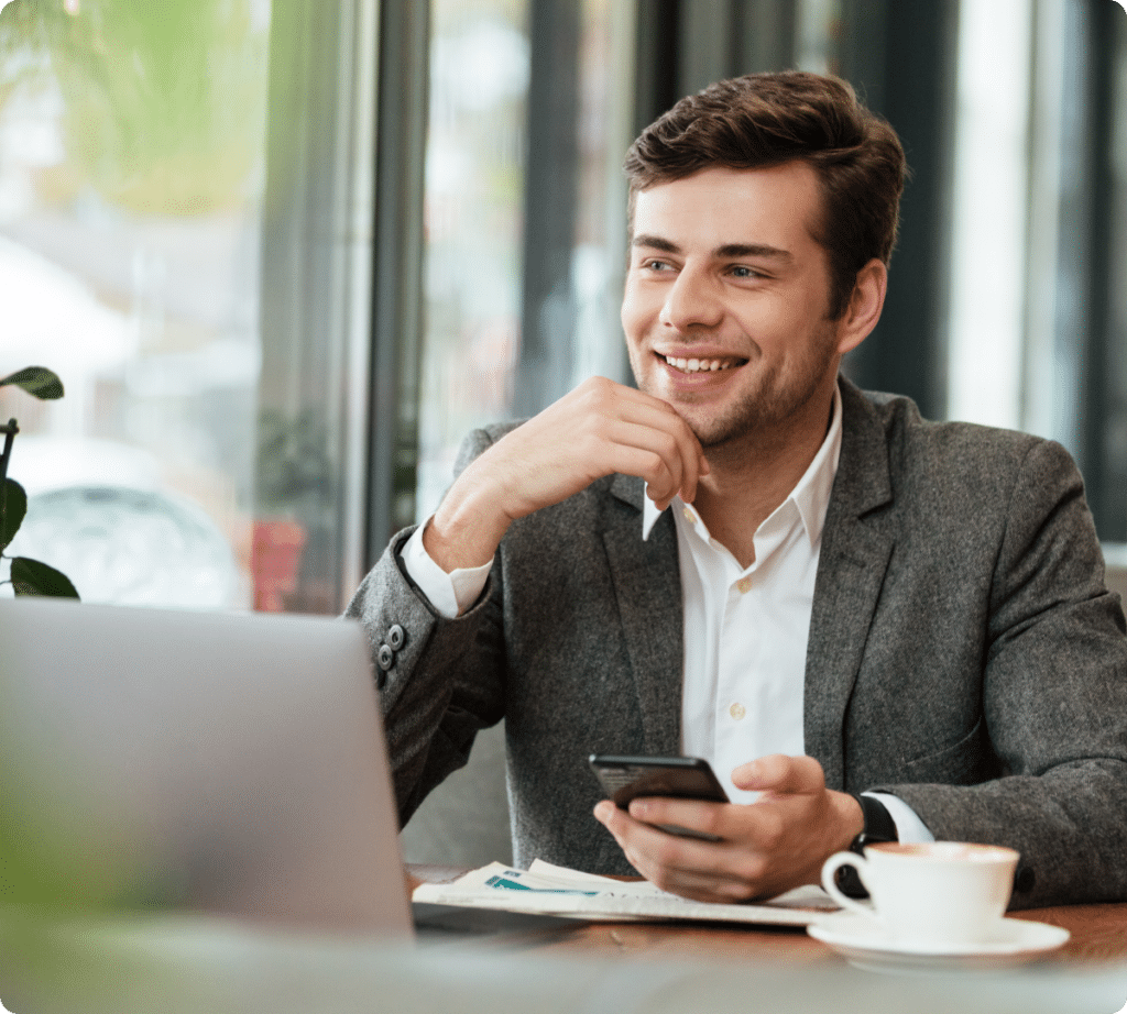 A man in a gray suit is smiling, holding a smartphone, and sitting at a table with a laptop and a cup of coffee in a bright cafe, possibly managing his ecommerce business.