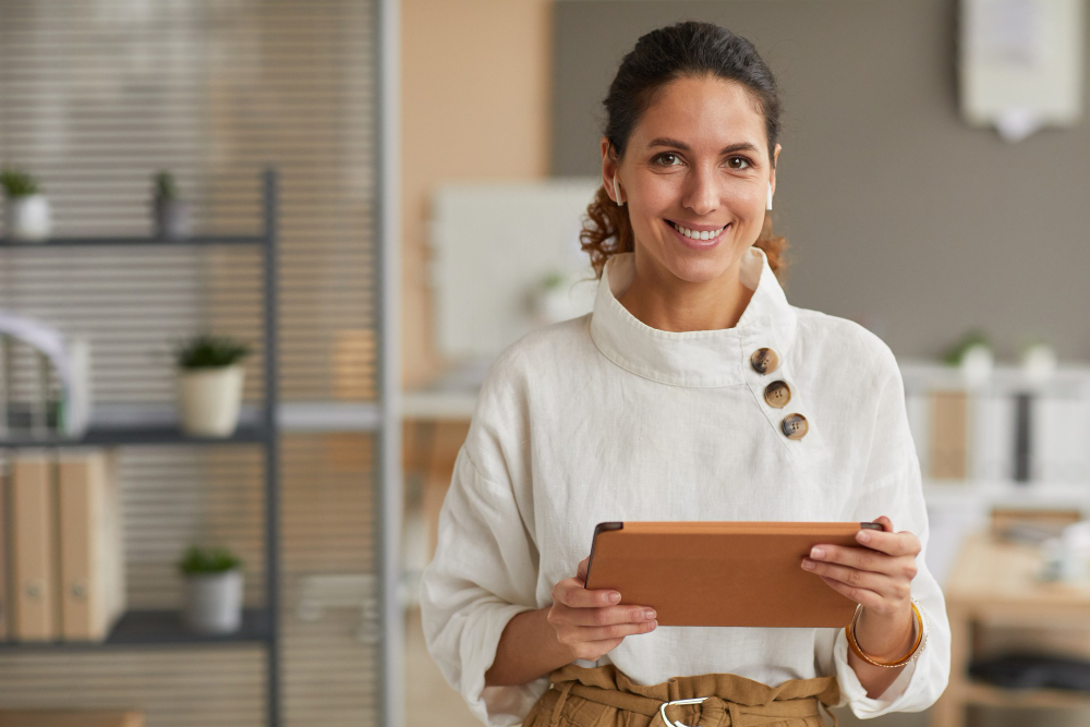 Woman in a white blouse holding a tablet, smiling and standing in an office environment with shelves and office supplies in the background, as she manages her ecommerce business.